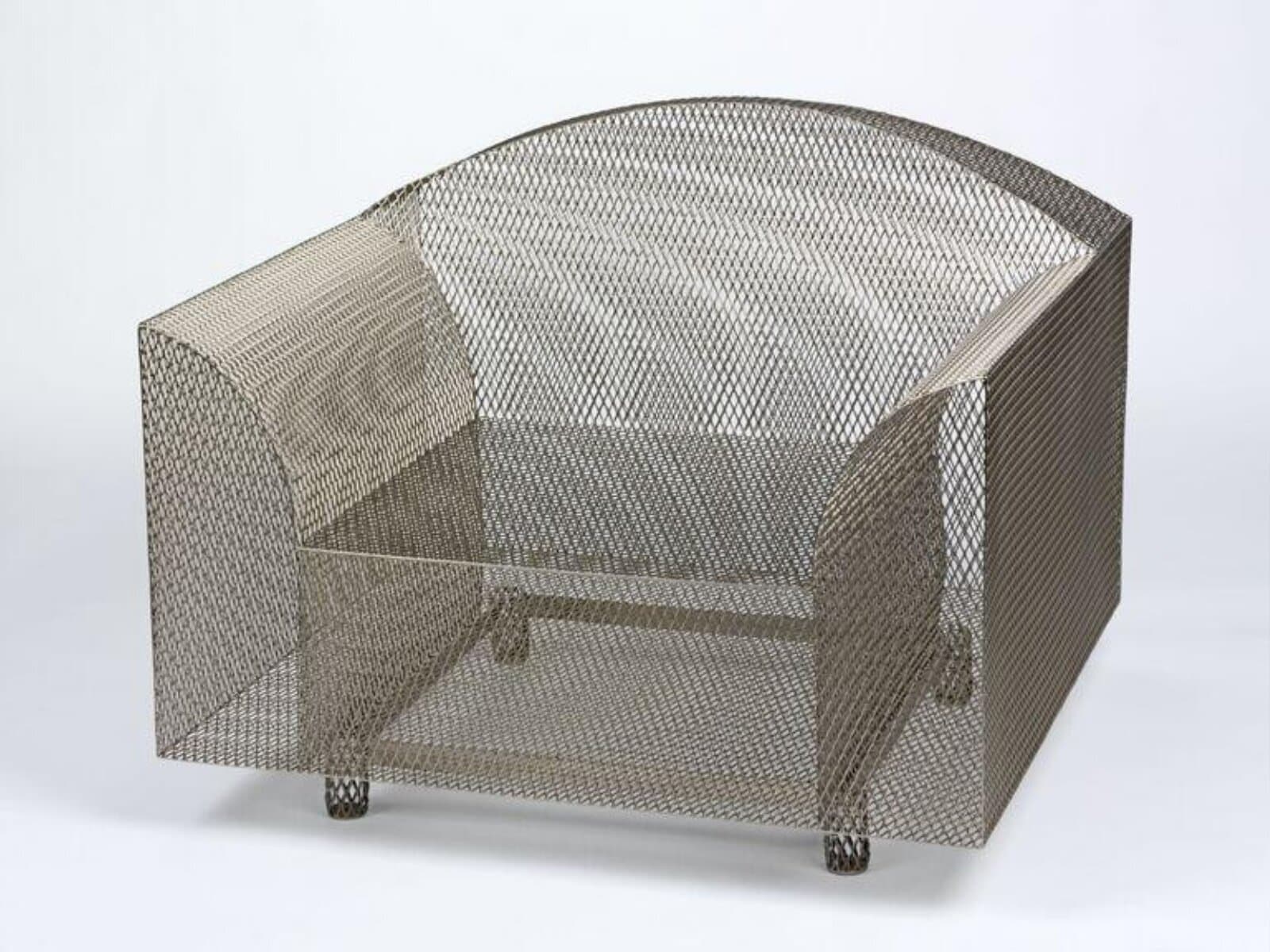 The mesh armchair at a three-quarter angle against a pale grey backdrop, showing the rounded top of the back, the flat side panels and four short cylindrical feet also made of mesh.