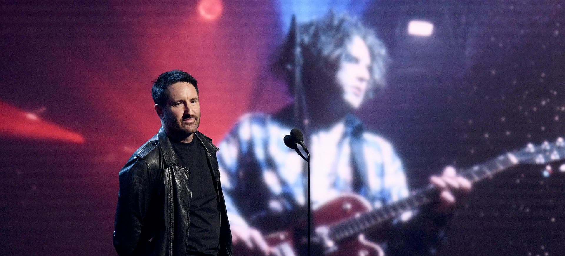 A man in a black leather jacket at a microphone on stage, a large screen behind him showing a guitarist.