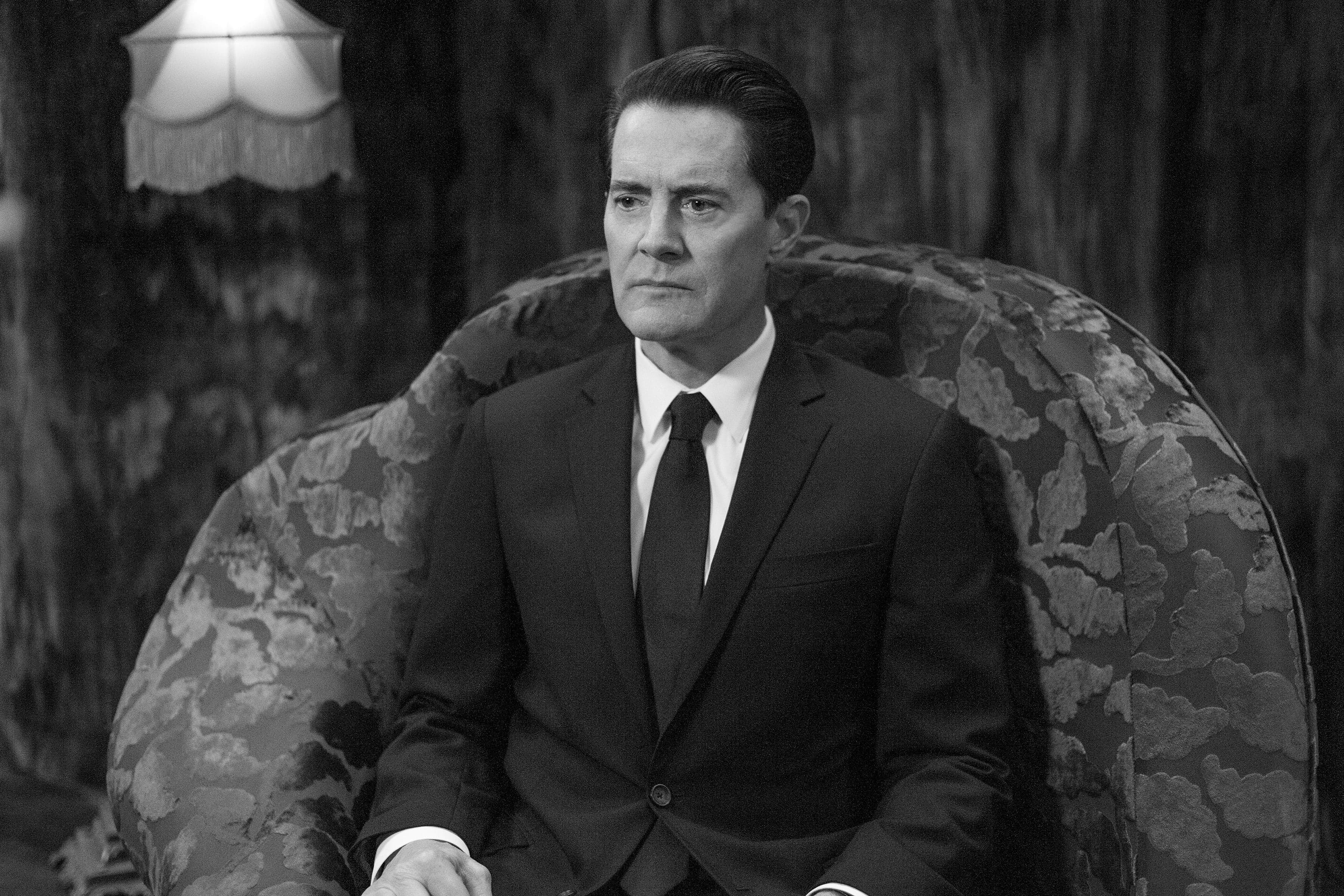 In black and white, Kyle MacLachlan in a dark suit sits upright in a large floral armchair under a fringed lamp shade.