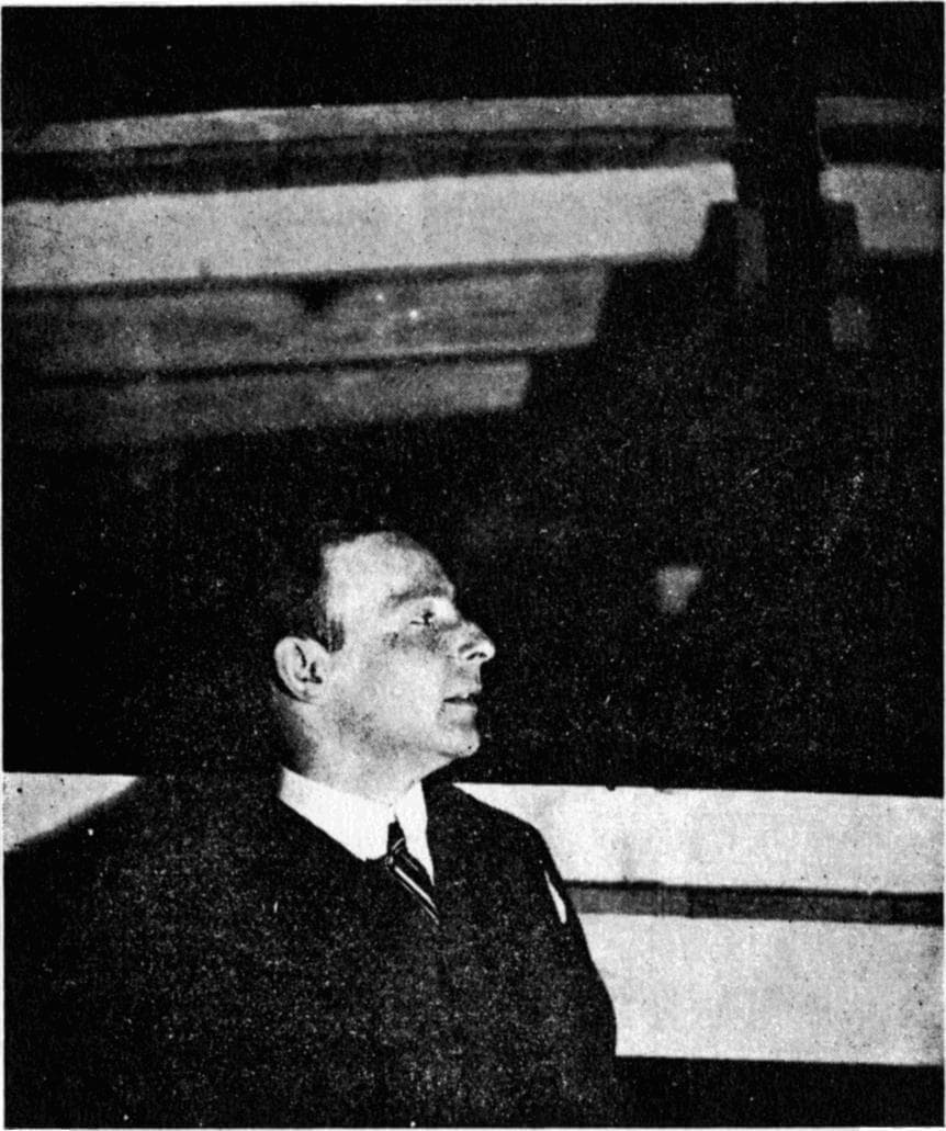 A black and white photograph of a man in a dark jacket and striped tie seen in profile, looking up at timber beams in a dim space.