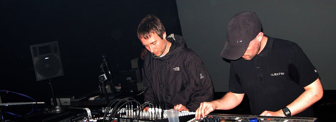 Two men lean over a table of mixers and cabled equipment in a dark room, one in a black jacket, one in a black cap and T-shirt.