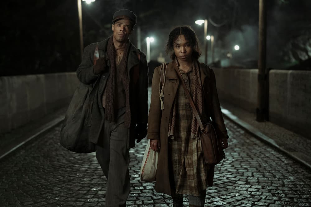 Jacob Anderson and Delainey Hayles, in worn wartime coats and carrying bags, walk toward the camera along a wet cobbled street at night.