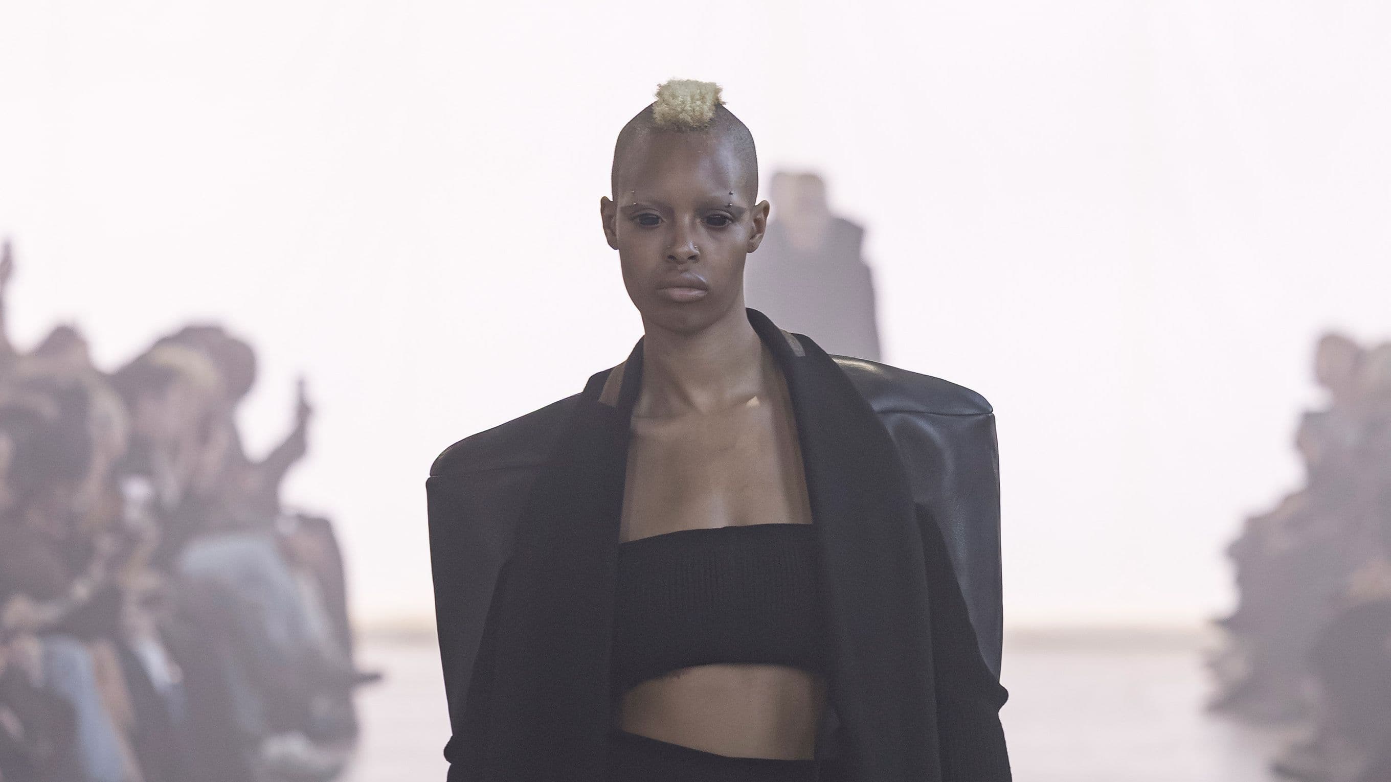 A model with a short blond crest walks the runway in a black bandeau and a black coat with huge squared shoulders.