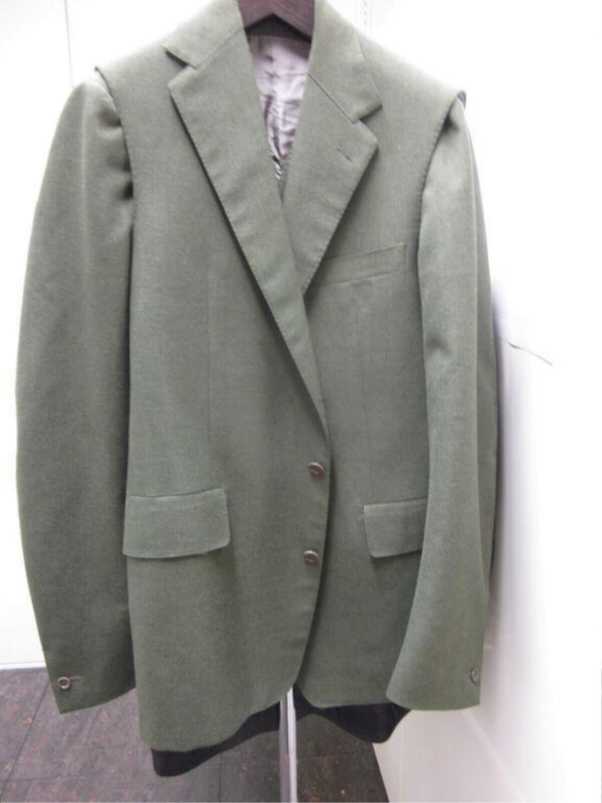 A khaki green tailored jacket on a hanger, shown full length, with the shoulder line of the sleeveless shell visible over the waistcoat's sleeves and a darker layer showing below the hem.