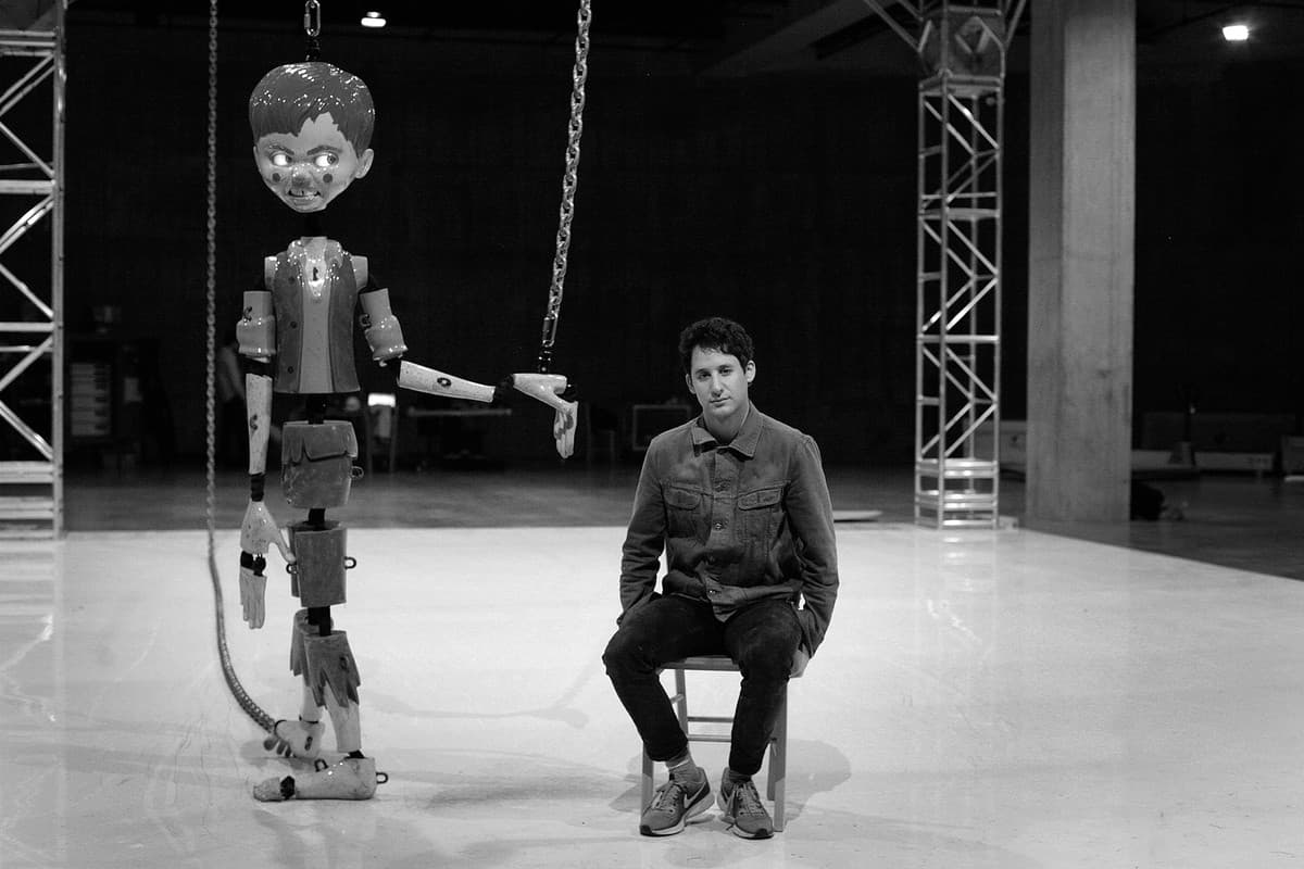 A man sits on a stool beside a suspended animatronic puppet on a bare stage rig, both lit hard against a dark industrial interior.