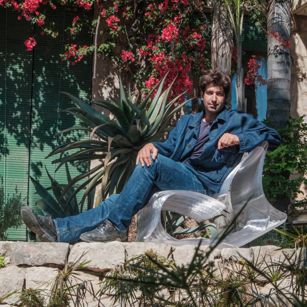 A man in denim sits in a clear sculpted chair on a stone ledge under bougainvillea, an agave beside him.