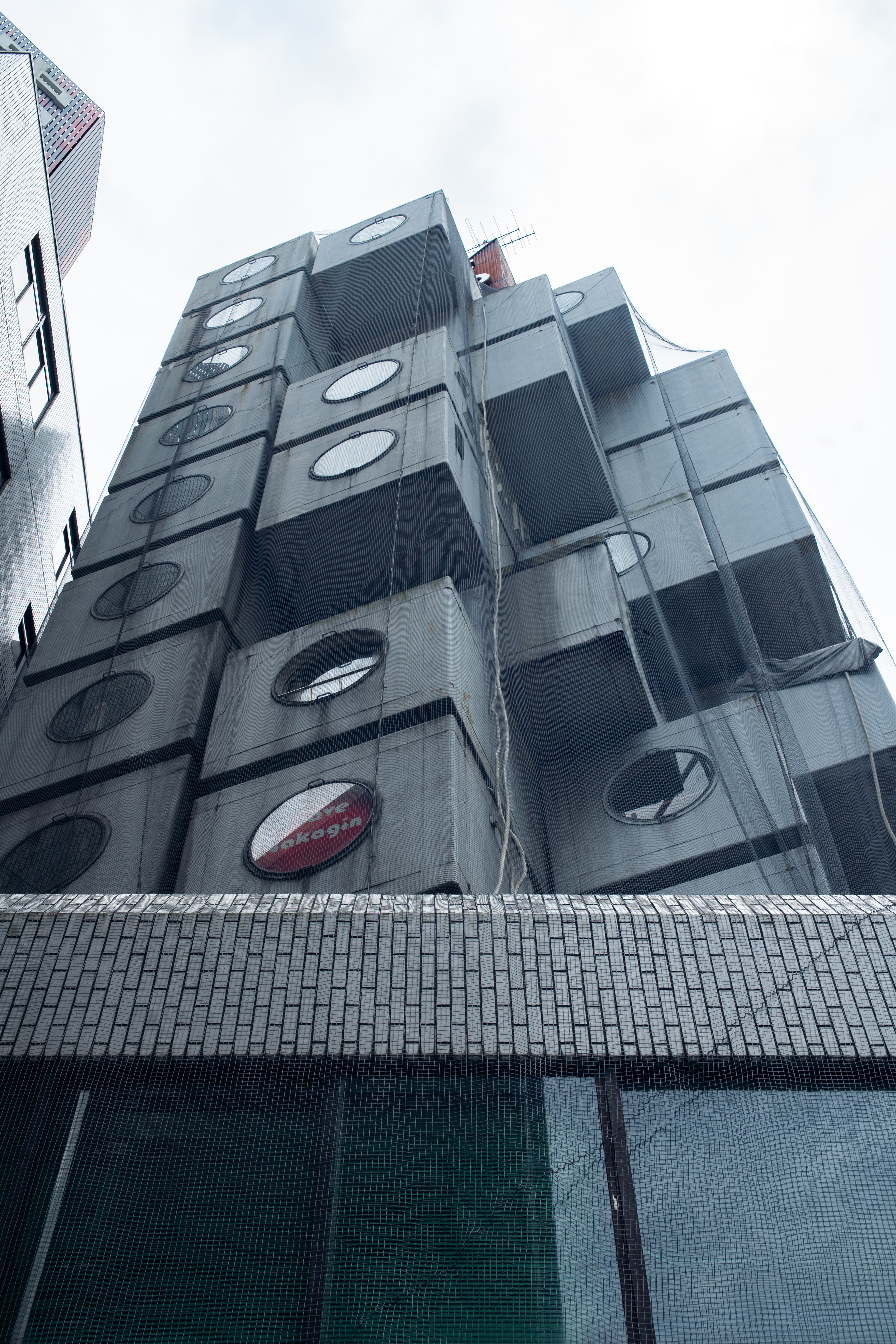 The Nakagin Capsule Tower seen from the street below, its stacked prefabricated capsules receding up the corner of the shaft against a flat grey sky.