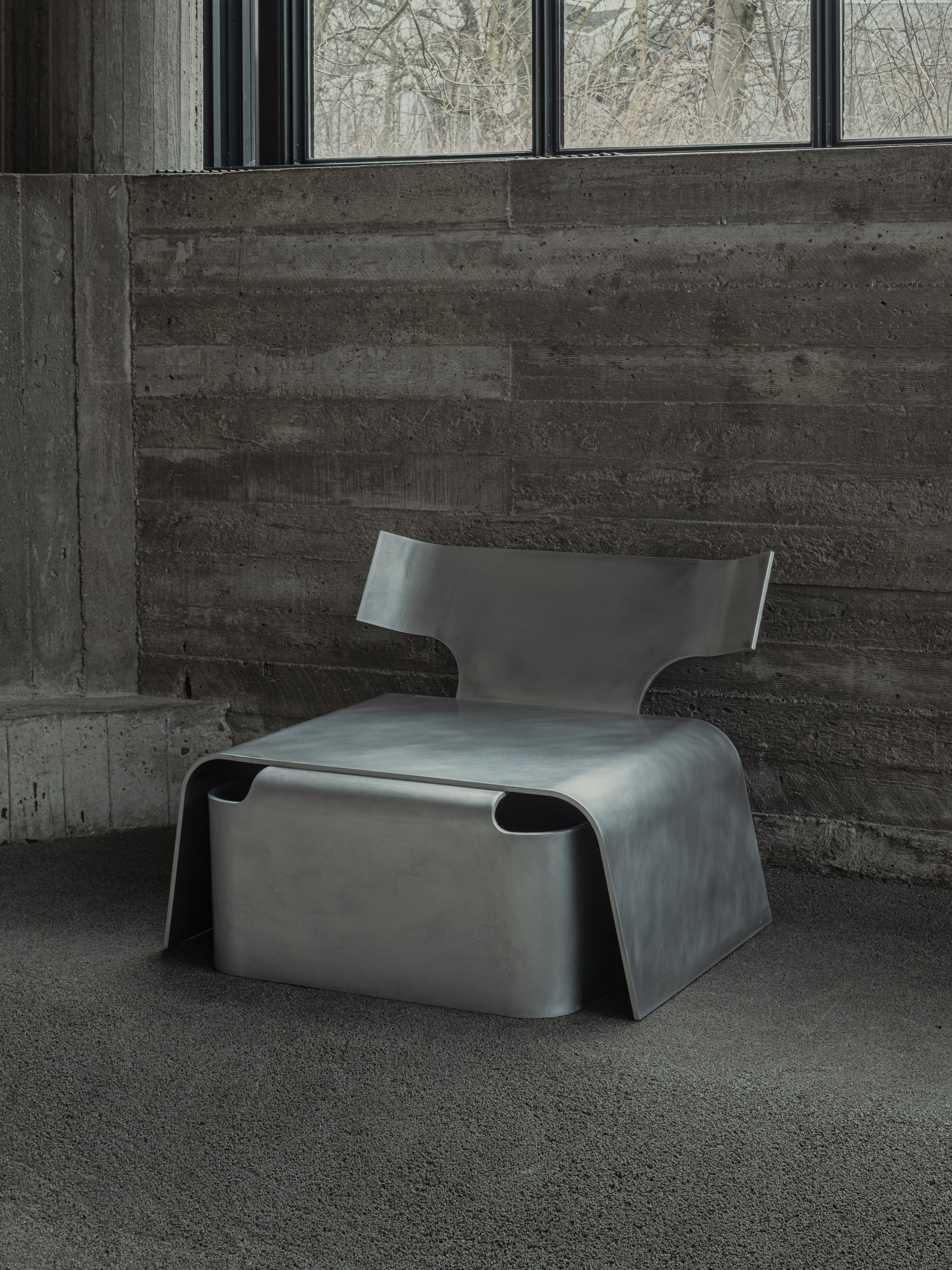 Linde Freya Tangelder's Low Chair in cast aluminium, a single angular seat standing against a board-formed concrete wall below a strip window.