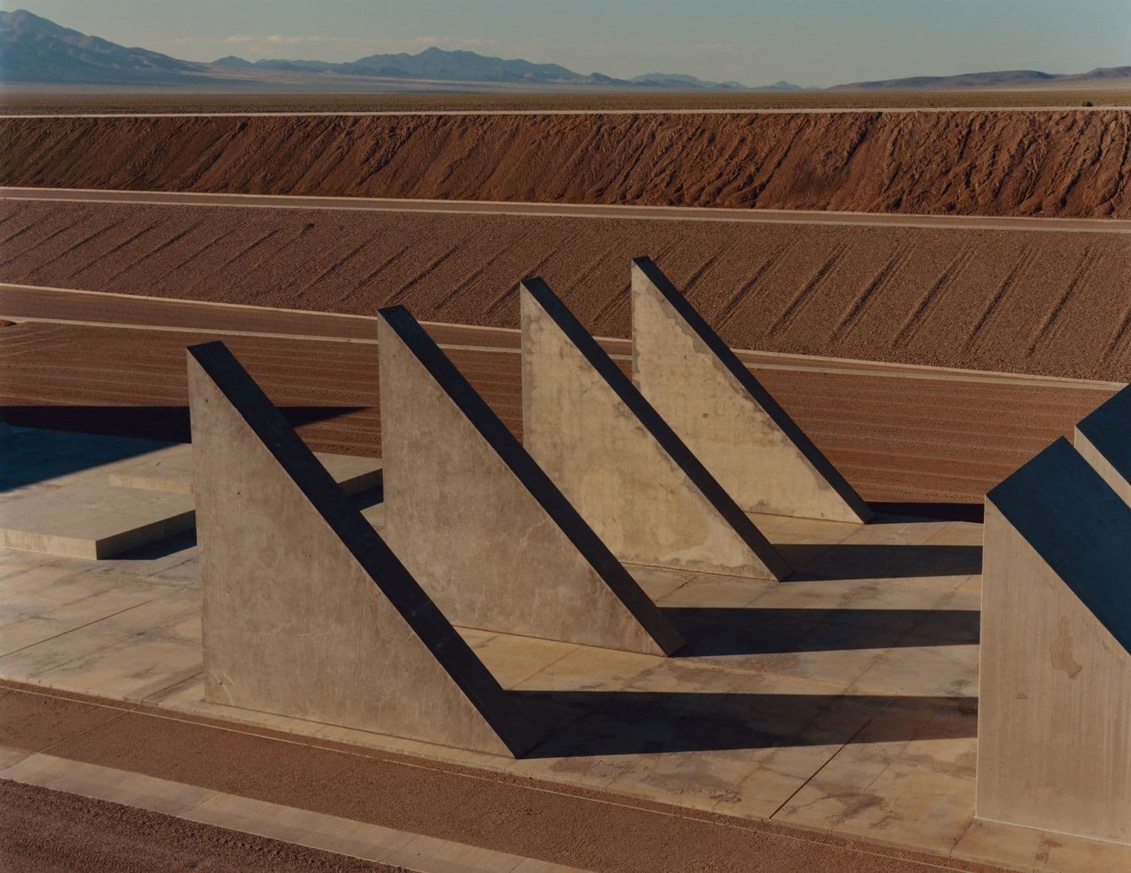 Rows of tall concrete wedge forms throw hard shadows across a pale concrete floor, with long earth berms and desert mountains behind.