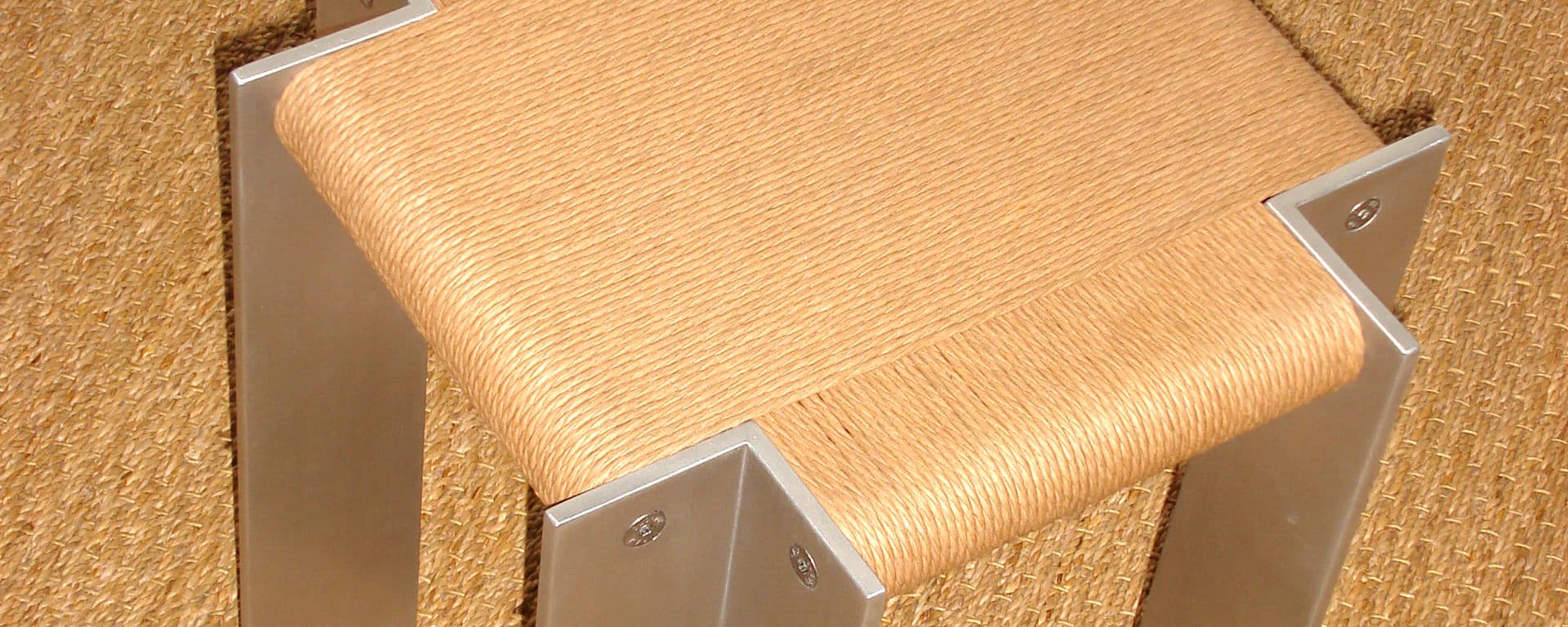 Close view of a stool seat wrapped in natural cord, held at the corners by brushed aluminium angle legs.