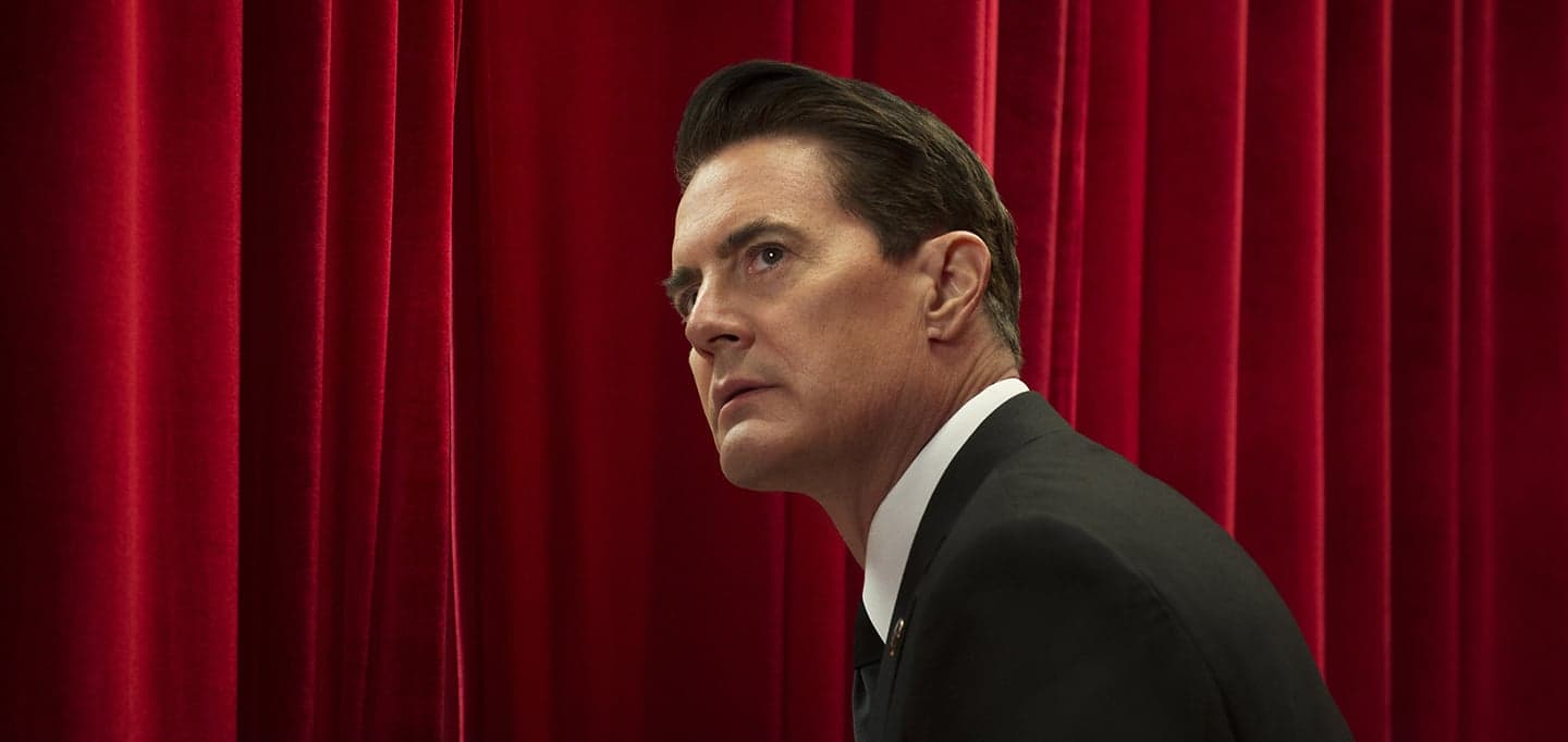 Kyle MacLachlan in a dark suit turns his head in front of a deep red velvet curtain.